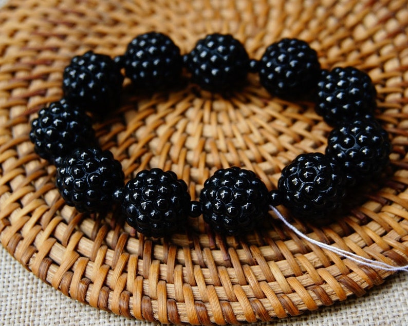 May include: A bracelet made of black, textured beads, arranged in a circular shape. The beads have a bumpy surface and are strung together with a white string. The bracelet is set against a woven, brown background.