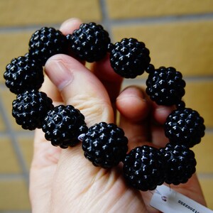 May include: A black beaded bracelet with textured, spherical beads. The bracelet is held in a hand, showcasing the glossy finish of the beads. The beads are strung together, forming a complete circle.