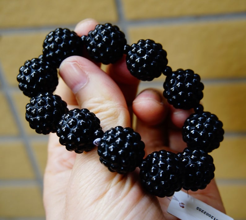 May include: A black beaded bracelet with textured, spherical beads. The bracelet is held in a hand, showcasing the glossy finish of the beads. The beads are strung together, forming a complete circle.
