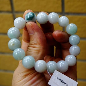 May include: A jade bracelet with large, spherical beads in pale green and white hues. The beads are strung together, forming a complete circle. The bracelet is held in a person's hand.
