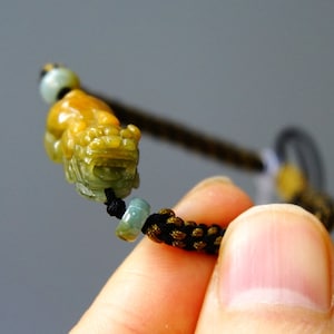 Könnte beinhalten: Ein Armband aus geflochtener Schnur in Schwarz und Braun mit einem Anhänger in Form eines Drachen aus Jadegrün und Gelb.