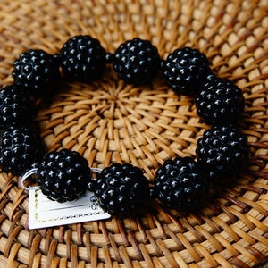 May include: A black beaded bracelet with a textured surface, resting on a woven wicker mat. The bracelet is composed of spherical beads, each with a detailed, bumpy texture. A small tag is attached to the bracelet.