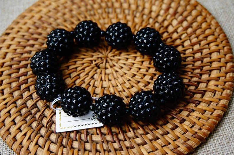 May include: A black beaded bracelet with a textured surface, resting on a woven wicker mat. The bracelet is composed of spherical beads, each with a detailed, bumpy texture. A small tag is attached to the bracelet.