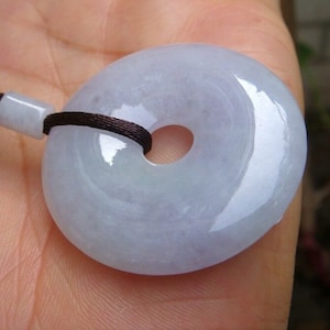 Op de afbeelding: Een lichtblauwe jade hanger in de vorm van een donut, met een gat in het midden. De hanger is aan een bruin koord.
