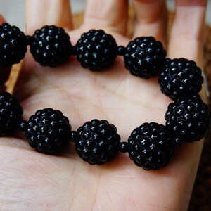 May include: A black beaded bracelet with a textured, spherical design. The bracelet is made of multiple round beads strung together, creating a bold and stylish accessory. The beads have a matte finish and are held in a hand.