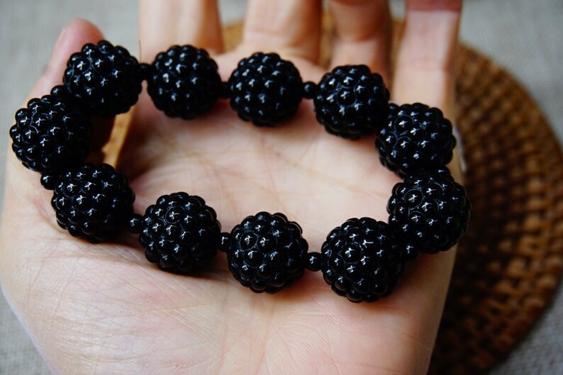 May include: A black beaded bracelet with a textured, spherical design. The bracelet is made of multiple round beads strung together, creating a bold and stylish accessory. The beads have a matte finish and are held in a hand.