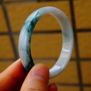 May include: A jade bangle bracelet with a smooth, polished surface. The bracelet features a blend of white and green hues, with the green colour concentrated in streaks. The bangle is held up against a blurred background.