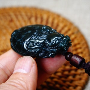 May include: A dark green jade pendant carved in the shape of a dragon. The pendant is attached to a dark brown cord with a black tassel.