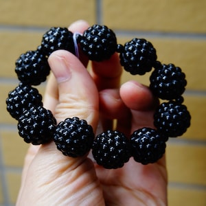 May include: A black beaded bracelet with textured, spherical beads. The bracelet is held in a hand, showcasing the glossy finish of the beads. The beads are strung together, forming a complete circle.