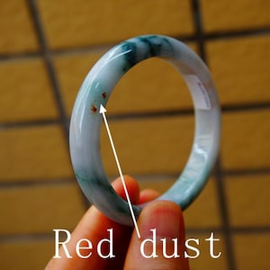 May include: A jade bangle bracelet with a smooth, polished surface. The bracelet features a blend of pale green and white hues, with darker green veining. Small red specks are visible on the surface. The bangle is held by a hand.