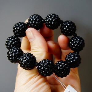 May include: A black beaded bracelet with a textured, spherical design. The bracelet is made of multiple round beads, each with a bumpy surface, strung together to form a circular shape. The beads are a glossy black color.