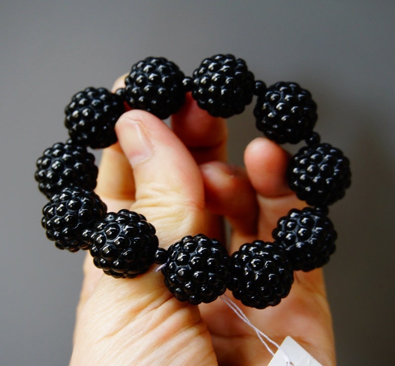 May include: A black beaded bracelet with a textured, spherical design. The bracelet is made of multiple round beads, each with a bumpy surface, strung together to form a circular shape. The beads are a glossy black color.