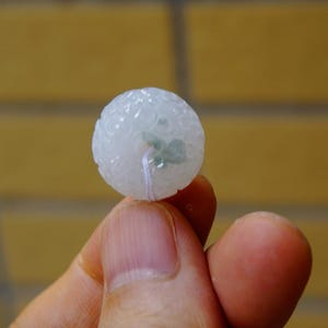May include: A round, translucent, white jade pendant with detailed floral carvings. The jade has a subtle green tint in the centre and is strung with a thin white cord. The pendant is held between two fingers.