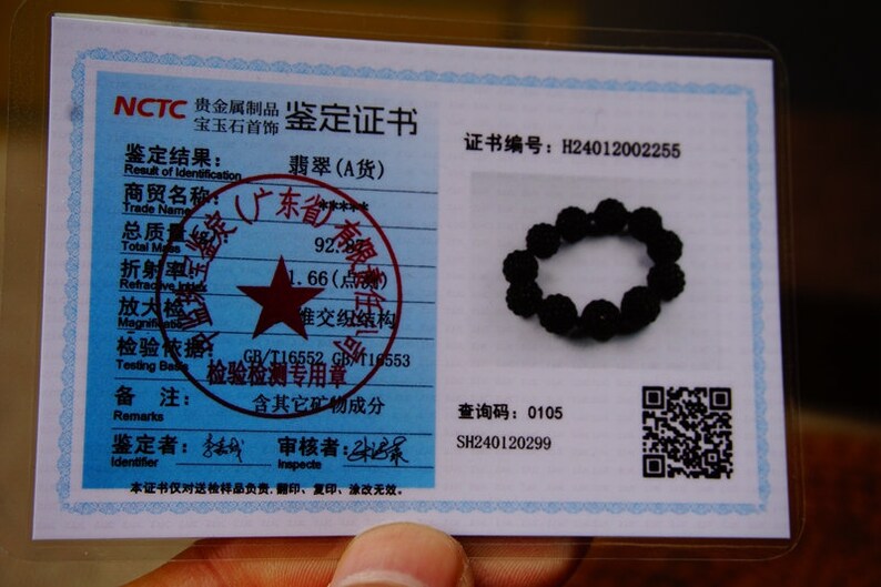 May include: A black beaded bracelet rests on a blue and white certificate. The certificate includes Chinese text, a red star, and a QR code. The bracelet is made of small, round, dark beads.