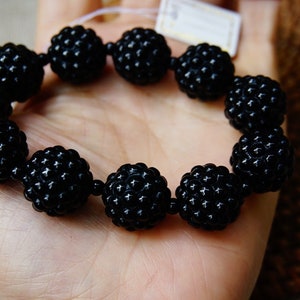 May include: A black beaded bracelet featuring large, round beads with a textured surface. The bracelet is held in a hand, showcasing its dark color and unique design. The beads appear to be approximately 1 inch in diameter.