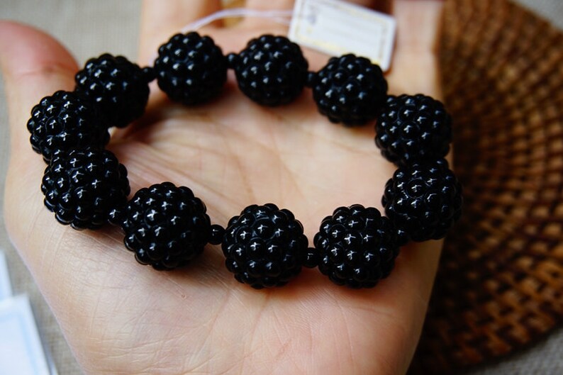 May include: A black beaded bracelet featuring large, round beads with a textured surface. The bracelet is held in a hand, showcasing its dark color and unique design. The beads appear to be approximately 1 inch in diameter.