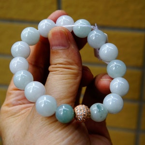 May include: A bracelet with large, round beads in white and pale green hues. One bead is rose gold and covered in sparkles, and two are a darker green. The bracelet is strung on a white cord.