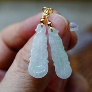 May include: A pair of jade feather earrings with gold accents. The earrings are carved in the shape of a feather and have a light green colour.