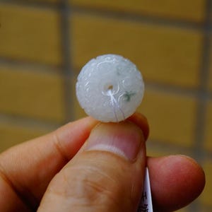 May include: A round, white jade bead with detailed floral carvings. The bead features a central hole and is strung on a thin thread. The jade has slight green markings.