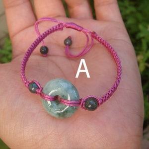 Peut inclure: Un bracelet en cordon tressé rose avec un pendentif en jade vert en forme de beignet et des perles noires. Le bracelet a une fermeture à nœud coulissant. La lettre "A" est écrite sur le bracelet.