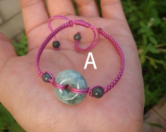 May include: A pink braided cord bracelet with a green jade donut charm and black beads. The bracelet has a sliding knot closure. The letter "A" is written on the bracelet.