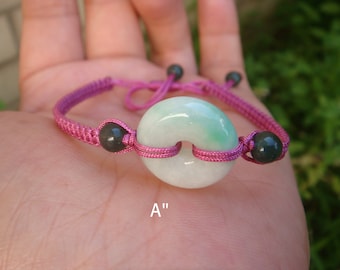 May include: A pink string bracelet with a green jade pendant in the shape of a circle with a hole in the middle. The pendant is about 1 inch in diameter. The bracelet is tied with a knot and has black beads on either side of the pendant.