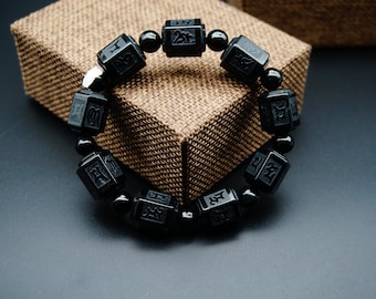 May include: Black obsidian bracelet with square beads featuring Chinese characters. The bracelet is on a brown textured surface.