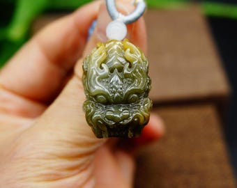 May include: A detailed close-up of a jade pendant carved in the shape of a mythical creature's head. The pendant is primarily olive green with yellow highlights, and is suspended from a light blue cord. The carving features intricate details.
