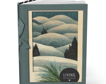 May include: A teal blue journal with a black and white patterned spine. The cover features a vintage illustration of a snowy landscape with trees and a small house. The text "LIVING OUR ADVENTURE" is printed in black on a white circle.