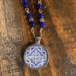 May include: A silver necklace with a round pendant featuring a blue and white tile design. The necklace has a chain of blue beads and silver accents, creating a vintage aesthetic.