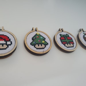 May include: Four small, round, wooden ornaments with white felt and colorful embroidery. The ornaments feature a mushroom with a Santa hat, a mushroom with a Christmas tree, a mushroom with a wrapped gift, and a mushroom with a Christmas pudding.