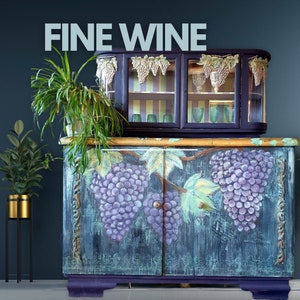 Könnte beinhalten: Ein dunkelblauer Schrank mit zwei Türen und Glasböden. Der Schrank ist mit einem bemalten Design aus Weinreben und Weintrauben verziert. Der Schrank hat einen goldenen Rahmen und die Worte "FINE WINE" sind in weißen Buchstaben obenauf geschrieben.