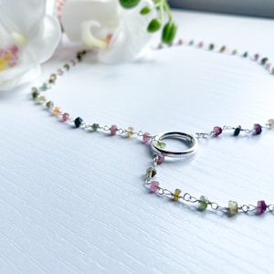 以下が含まれることがあります： 円形のペンダントと小さなカラフルな宝石が付いたシルバーネックレス。ネックレスは白い表面の上に置かれています。