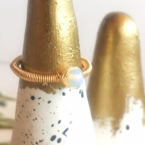 Puede incluir: Un anillo de alambre dorado con una piedra de &oacute;palo blanco. El anillo se muestra en un soporte de anillo blanco y dorado.