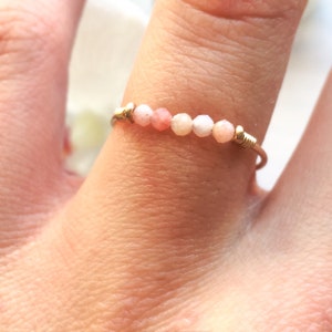 Pu&ograve; includere: Un anello delicato con piccole perline rosa e bianche sfaccettate infilate su un sottile filo d'oro. Le perline sono disposte in fila sulla parte superiore dell'anello, creando un design minimalista ed elegante. L'anello &egrave; mostrato su un dito.