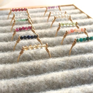 Pu&ograve; includere: Una collezione di delicati anelli dorati, ciascuno con una fila di pietre preziose colorate. Gli anelli sono esposti su una superficie grigia foderata di feltro. Le pietre preziose includono rosso, blu, giallo, verde e rosa.