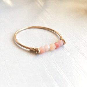 Pu&ograve; includere: Un delicato anello color oro con una fila di perline sfaccettate rosa e pesca. Le perline sono infilate su un filo sottile, creando un design minimalista ed elegante. L'anello &egrave; un gioiello semplice ma elegante.