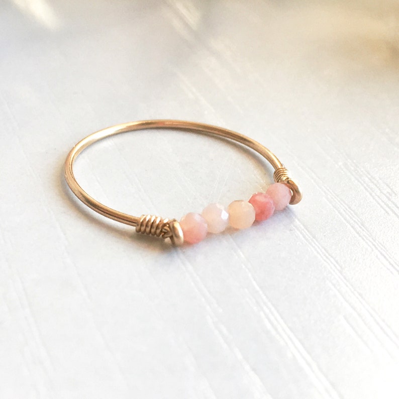 Pu&ograve; includere: Un delicato anello color oro con una fila di perline sfaccettate rosa e pesca. Le perline sono infilate su un filo sottile, creando un design minimalista ed elegante. L'anello &egrave; un gioiello semplice ma elegante.