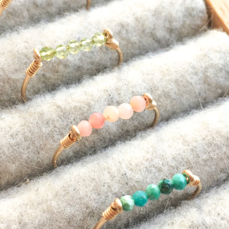 Pu&ograve; includere: Primo piano di tre anelli fatti a mano con filo color oro e perline di pietre preziose colorate. Un anello presenta perline verdi, un altro ha perline rosa e il terzo perline turchesi. Gli anelli sono esposti su una superficie grigio chiaro.