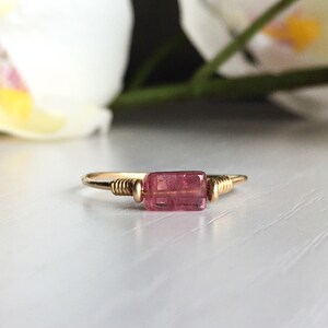 May include: A delicate gold ring featuring a pink tourmaline gemstone. The ring is crafted with a simple wire design and showcases a rectangular-shaped stone.
