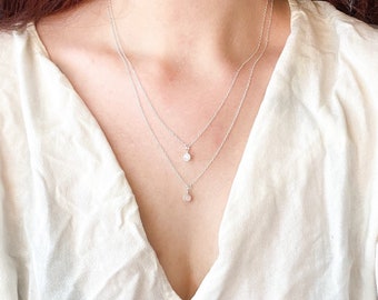 Peut inclure: Un collier en chaîne argent avec deux pendentifs. Le pendentif supérieur est une petite pierre ronde et transparente. Le pendentif inférieur est une pierre ronde opaque plus grande.