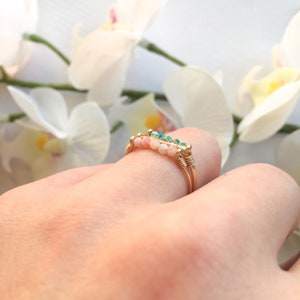 Pu&ograve; includere: Un anello in filo dorato con file di piccole perline rotonde. Le perline sono un mix di rosa e turchese, creando un accento colorato. L'anello &egrave; mostrato su un dito, con fiori bianchi sullo sfondo.