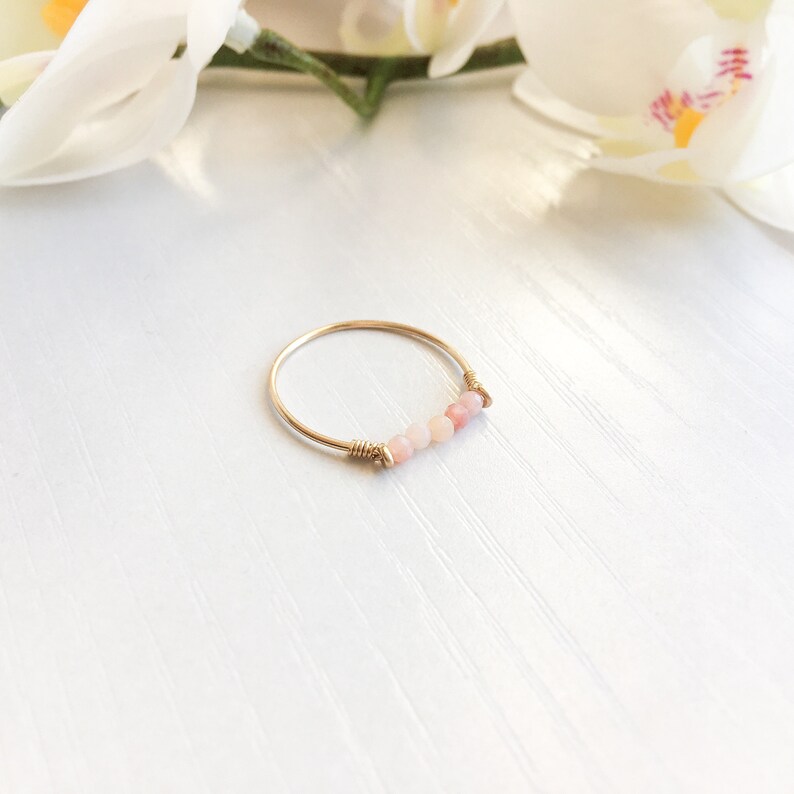 Pu&ograve; includere: Un delicato anello d'oro con una fila di piccole pietre preziose rosa chiaro. L'anello ha una sottile fascia dorata, con le pietre preziose incastonate al centro. Le pietre preziose sono di un delicato colore rosa, aggiungendo un tocco di colore.