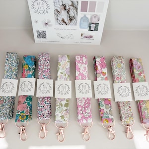 May include: An assortment of floral lanyards with rose gold clasps. Each lanyard displays a unique, colourful floral pattern, with a white tag bearing the text "Wild Ivy". The lanyards are neatly arranged, highlighting the range of designs available.