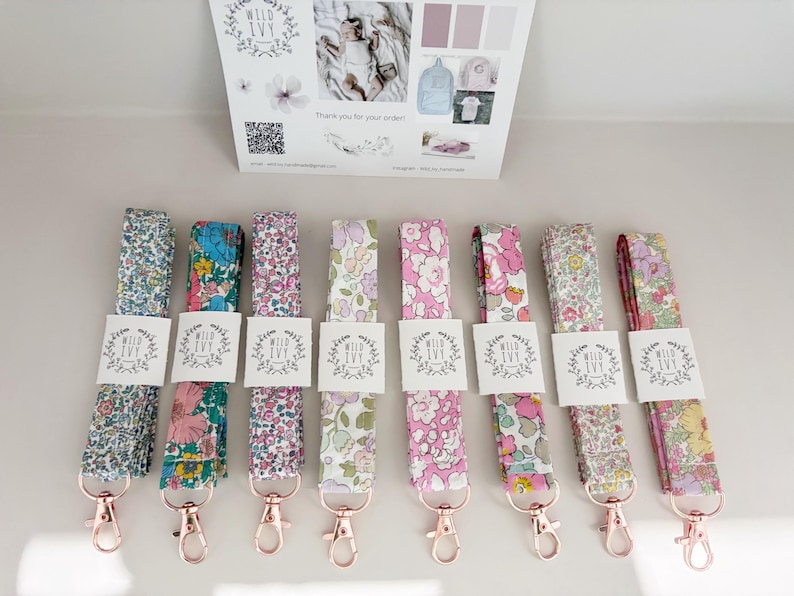 May include: An assortment of floral lanyards with rose gold clasps. Each lanyard displays a unique, colourful floral pattern, with a white tag bearing the text "Wild Ivy". The lanyards are neatly arranged, highlighting the range of designs available.