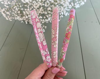 May include: Three pink fabric headbands with floral patterns. One features white cartoon-like figures, another has a delicate floral print, and the third showcases pink and red flowers. The headbands are held in front of a blurred background of white flowers.