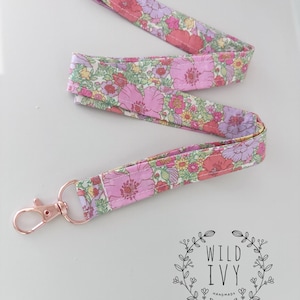 May include: A floral patterned lanyard with a rose gold coloured clasp. The fabric displays pink, purple, and yellow flowers on a white background. The lanyard is made of a soft, flexible material.