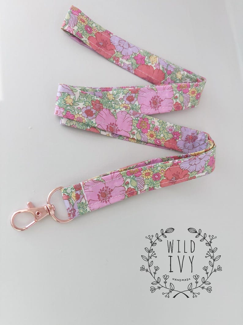 May include: A floral patterned lanyard with a rose gold coloured clasp. The fabric displays pink, purple, and yellow flowers on a white background. The lanyard is made of a soft, flexible material.