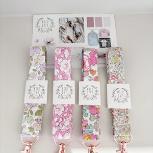 May include: Four floral lanyards with rose gold clasps. Each lanyard features a different floral design in pink, purple, and yellow hues. Each lanyard has a white tag with the text "WILD IVY". A card with the "WILD IVY" logo and baby photos is in the background.