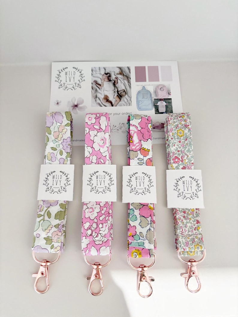 May include: Four floral lanyards with rose gold clasps. Each lanyard features a different floral design in pink, purple, and yellow hues. Each lanyard has a white tag with the text "WILD IVY". A card with the "WILD IVY" logo and baby photos is in the background.